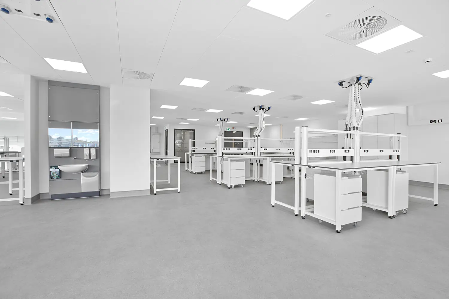 Spacious, modern laboratory fitted with white workbenches arranged across a wide, clean grey floor. Each bench features integrated storage drawers and overhead service units suspended from the ceiling, equipped with flexible hoses and power or data outlets. The ceiling is lined with recessed square light panels and circular ventilation grilles, creating an even, bright illumination throughout the room.  On the left, a recessed wash station with a sink, mirror and grey panel detailing sits beside a column, while the rest of the space extends open and uncluttered towards the back, where doors and additional work areas are visible. The overall setting is minimal, clinical and highly organised, with a focus on functionality and clean lines.