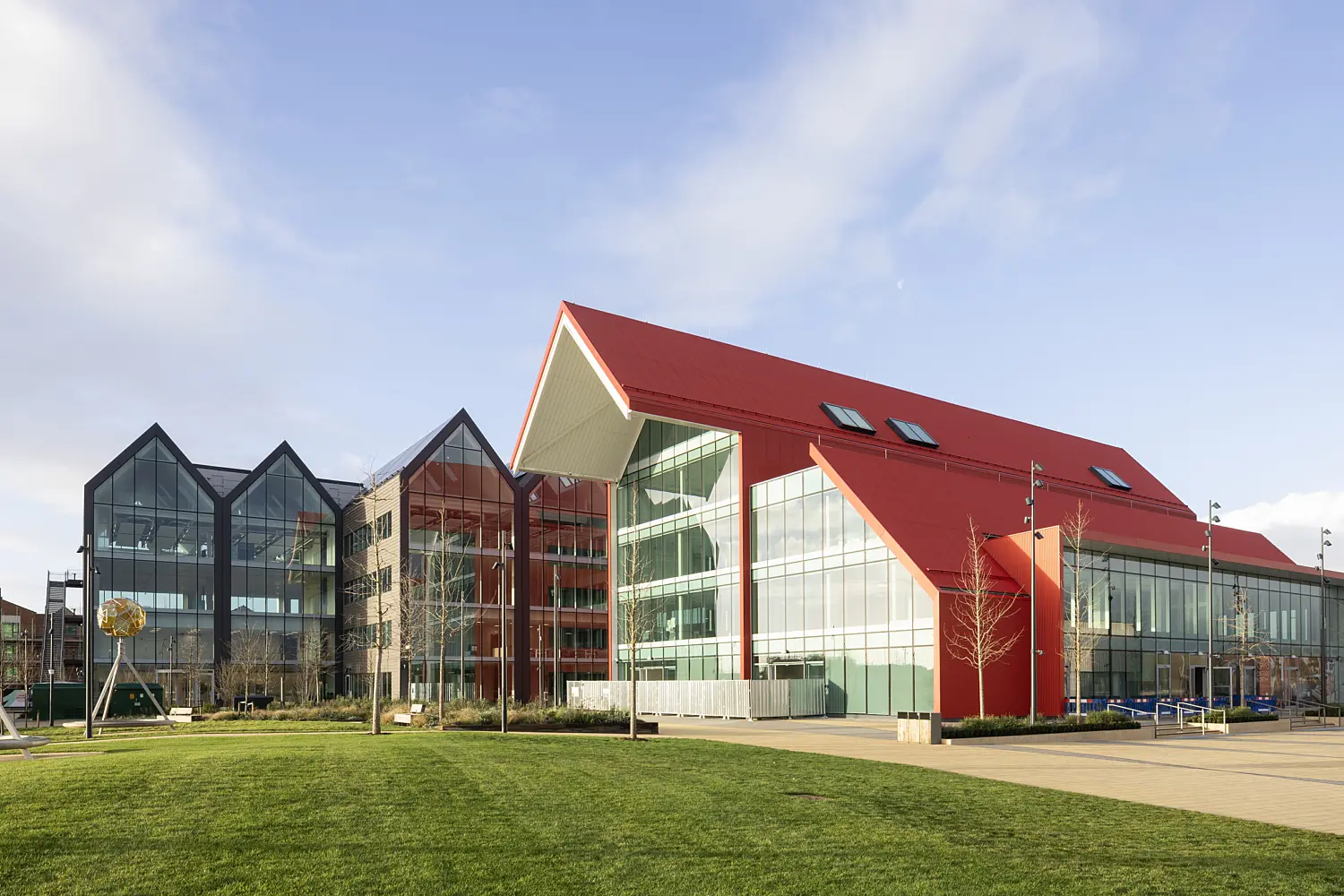 Modern campus-style building with a red pitched roof and large glass façades, connected to a darker grey, multi-gabled glass structure on the left. The buildings sit beside a wide paved walkway and a neatly maintained green lawn in the foreground. Sculptural metal frames holding spherical artworks stand on the grass. The scene is brightly lit by sunlight under a clear blue sky with light clouds, highlighting the clean lines, reflective windows and contemporary architectural design.