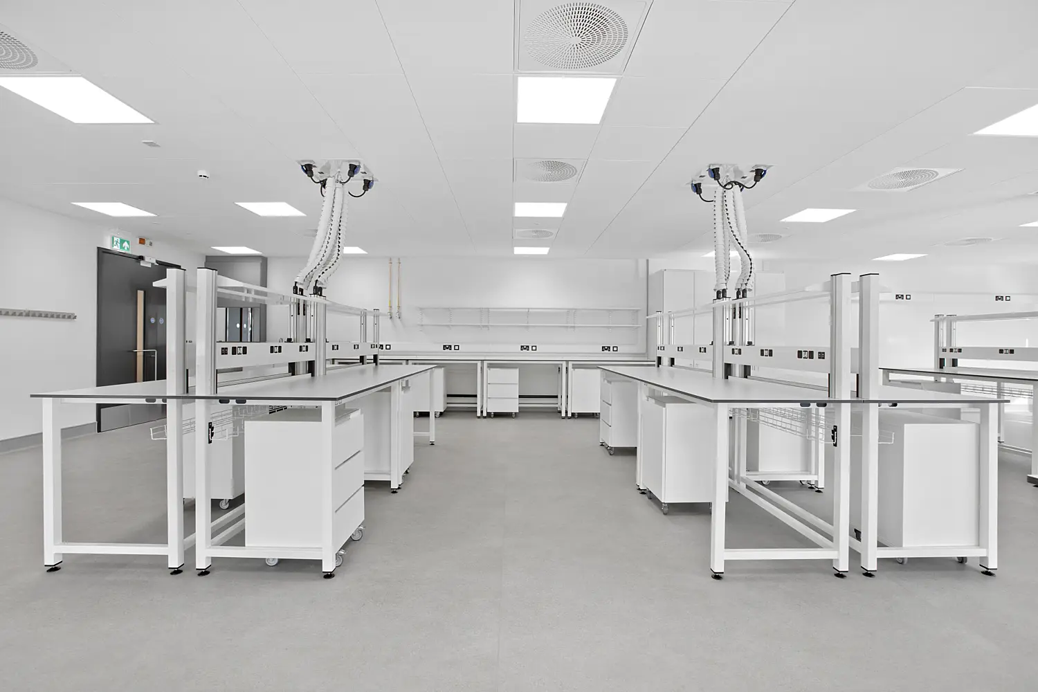 Clean, contemporary laboratory interior with two rows of white workbenches facing each other across a central aisle. Each bench includes integrated shelving and suspended ceiling service units with flexible hoses and multiple outlets. The room features a smooth grey floor, white ceiling panels with recessed square lights and circular ventilation grilles, and built-in cabinetry and shelving along the back wall. A dark door with a green emergency exit sign is visible on the left, while the overall space appears bright, orderly and uncluttered with a clinical, modern finish.