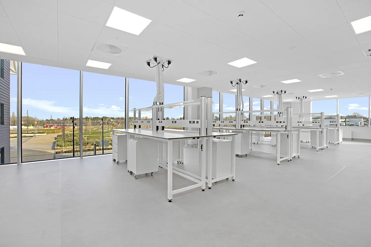 Bright, modern laboratory or classroom space with white workbenches arranged in rows on a smooth grey floor. Each bench is fitted with overhead service units suspended from the ceiling, featuring multiple outlets and flexible connections. Large floor-to-ceiling windows line one side of the room, allowing natural light to flood the space and offering views of landscaped grounds outside. The ceiling is fitted with recessed square lighting panels and ventilation fixtures, reinforcing the clean, clinical environment.