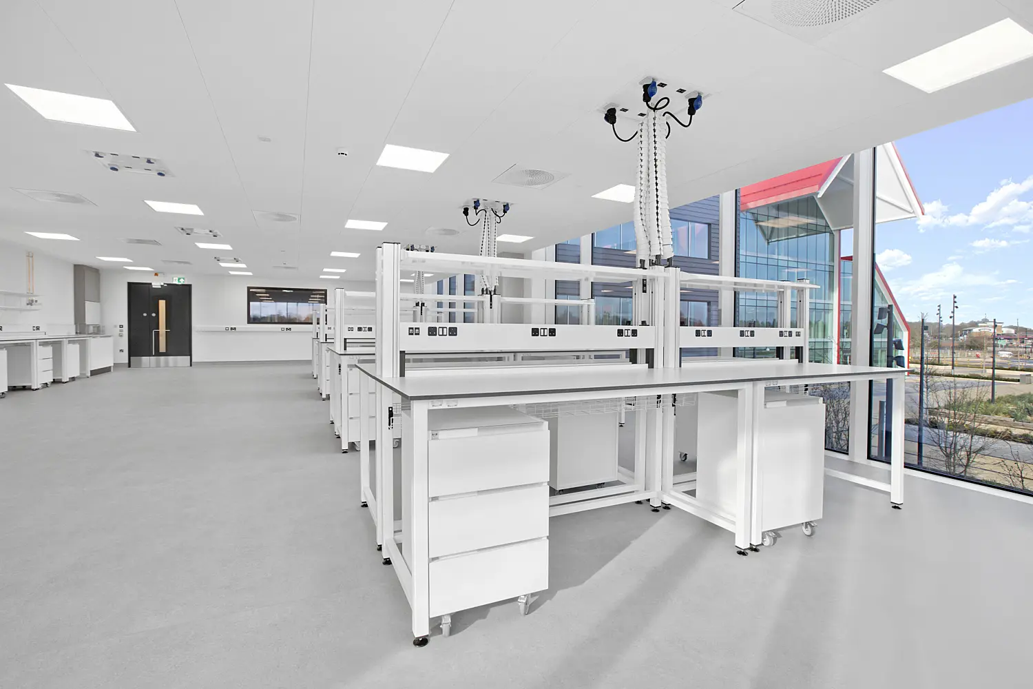 Spacious, modern laboratory with rows of white workbenches fitted with integrated shelving and suspended service units overhead. Each unit features multiple outlets and flexible connections, positioned above the benches for easy access. Large floor-to-ceiling windows run along the right side, filling the room with natural light and revealing views of landscaped grounds and a contemporary red-roofed building outside. The interior has a clean, minimal aesthetic with a grey floor, white ceiling panels with recessed lighting and ventilation fixtures, and additional cabinetry and equipment lining the far wall.
