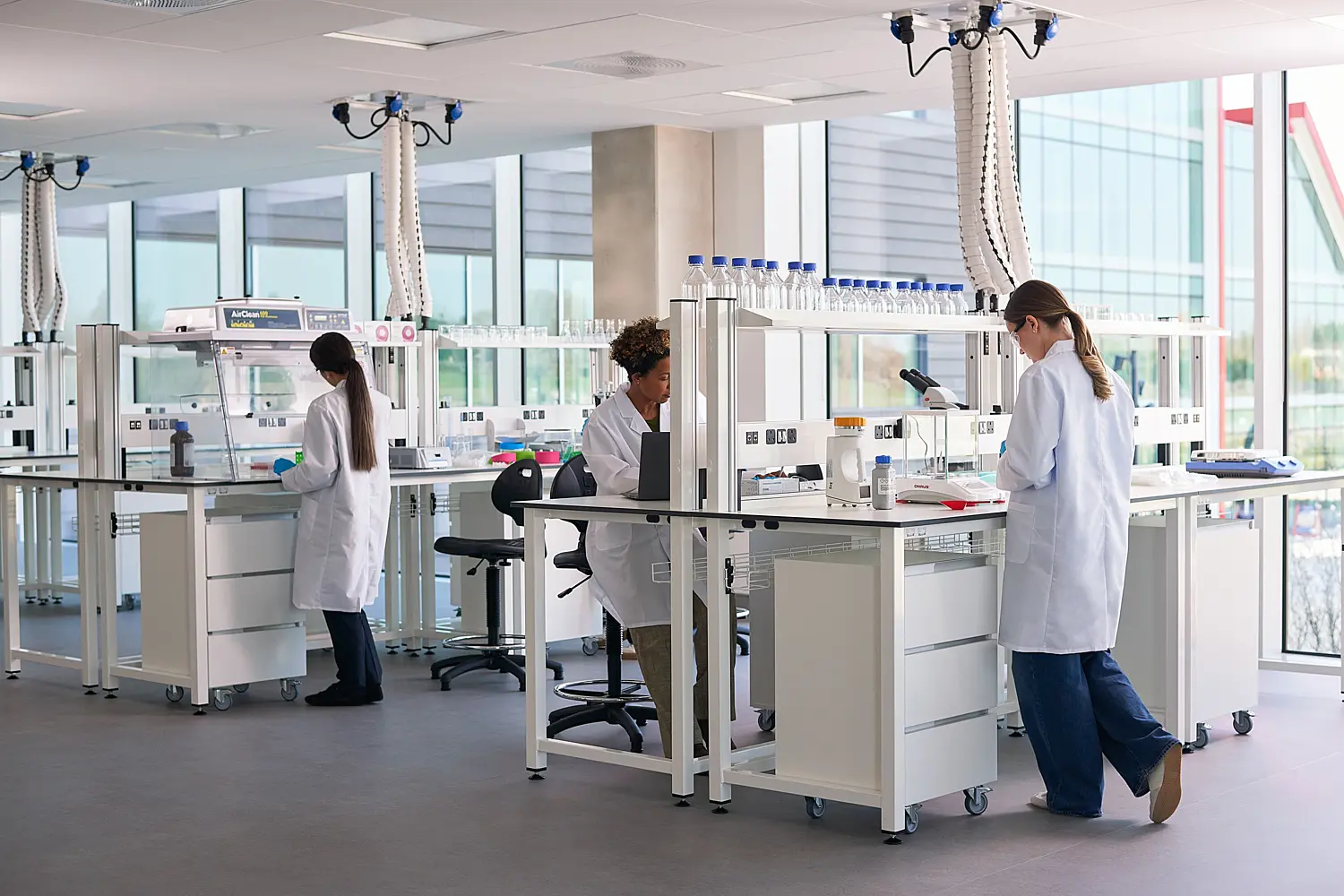 Bright, modern laboratory with large floor-to-ceiling windows, white workstations and scientific equipment. Three researchers in white lab coats work at separate benches, using laboratory instruments and computers. Shelving holds rows of sample bottles and glassware, while ceiling-mounted extraction arms hang above the work areas. The space is clean, open and filled with natural daylight.