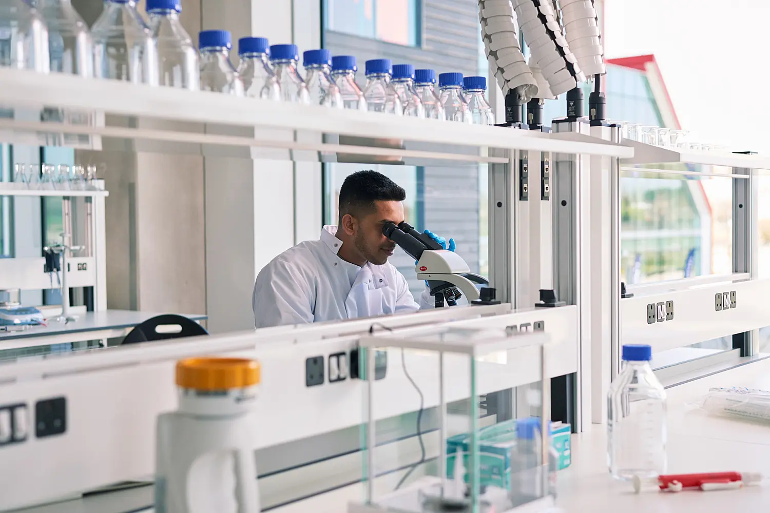 Research laboratory with white benches, glassware and rows of sample bottles on shelving. A laboratory worker in a white coat uses a microscope at a workstation surrounded by scientific equipment. Large windows fill the space with natural light, while ceiling-mounted extraction arms and clean, modern fittings create a contemporary research environment.