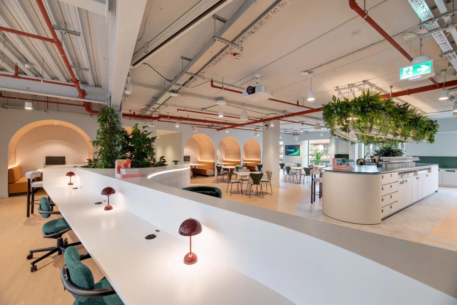 A spacious office with long white workbenches in the foreground, each fitted with small dome-shaped desk lamps and green task chairs. In the centre of the space, arched alcoves with warm lighting create semi-enclosed seating areas. To the right, a large curved counter sits beneath a ceiling feature covered with hanging plants. Exposed services run across the ceiling, and the setting combines planted areas, soft seating and communal tables.