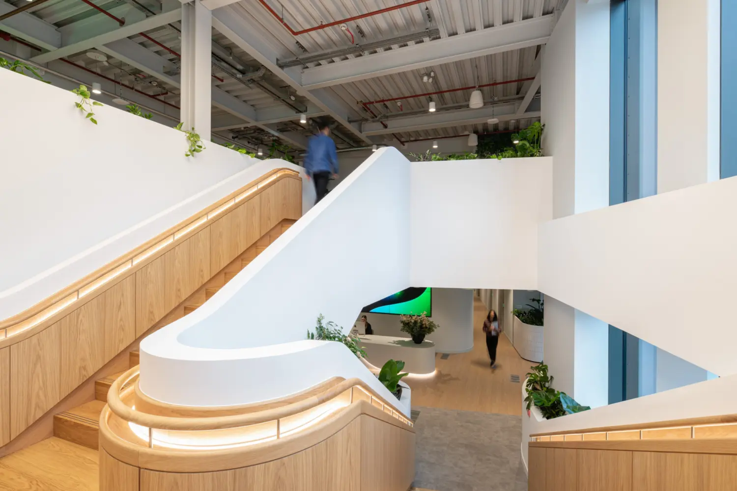 A modern stairwell with white balustrades and timber accents, featuring integrated lighting along the handrails. The staircase splits across multiple levels, with planted areas at the landings. Exposed ceiling services run above, and large windows on the right bring natural light into the space. People are visible moving through the area, giving a sense of scale and activity.