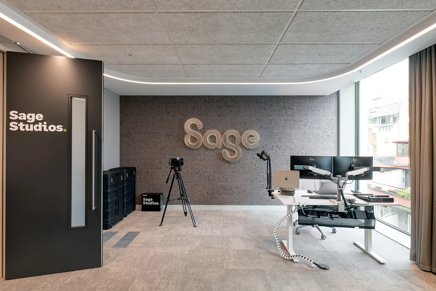 A modern studio space with a textured feature wall displaying the Sage logo. To the left is a black door marked “Sage Studios”, and a camera on a tripod is set up in front of the wall. On the right, a desk with multiple monitors and equipment sits beside full-height windows that look out onto neighbouring buildings. The room has soft grey flooring, a suspended ceiling and clean, contemporary finishes.