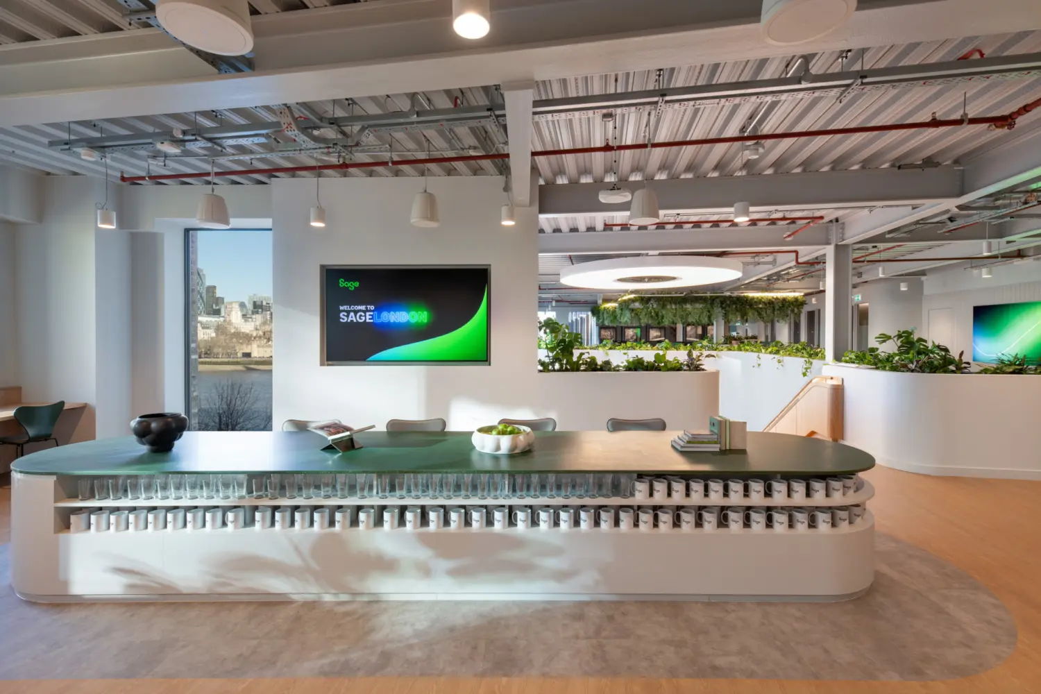 A bright office space with a long green-topped counter in the foreground, backed by neatly arranged rows of cups. A large wall screen is mounted to the left, and daylight enters through a window behind the desk. Beyond the counter, planted partitions frame an open workspace with exposed services, soft lighting and a circular ceiling feature above a greenery-filled area. A staircase with timber treads is visible on the right.