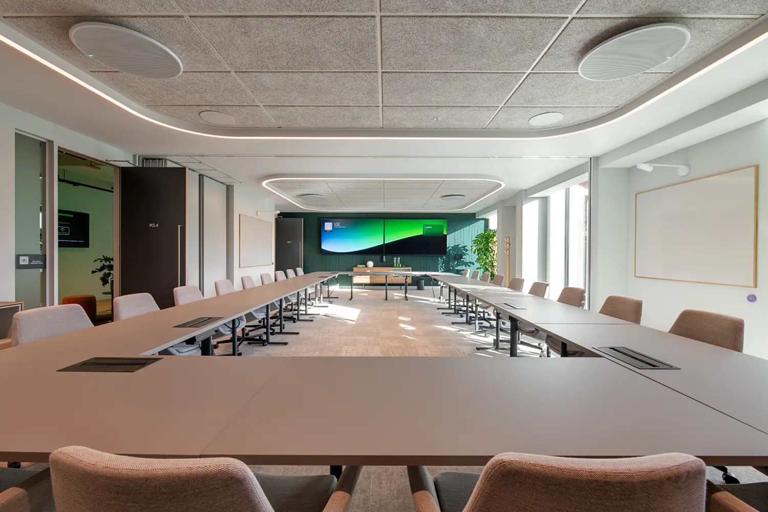 A large meeting room with two long tables arranged in a U‑shape, each surrounded by upholstered chairs. A wide digital screen spans the front wall, with a small seating area positioned below it. Floor‑to‑ceiling windows on the right bring in natural light. The ceiling features acoustic panels with integrated lighting, and the room is finished in soft neutral tones with minimal decoration.