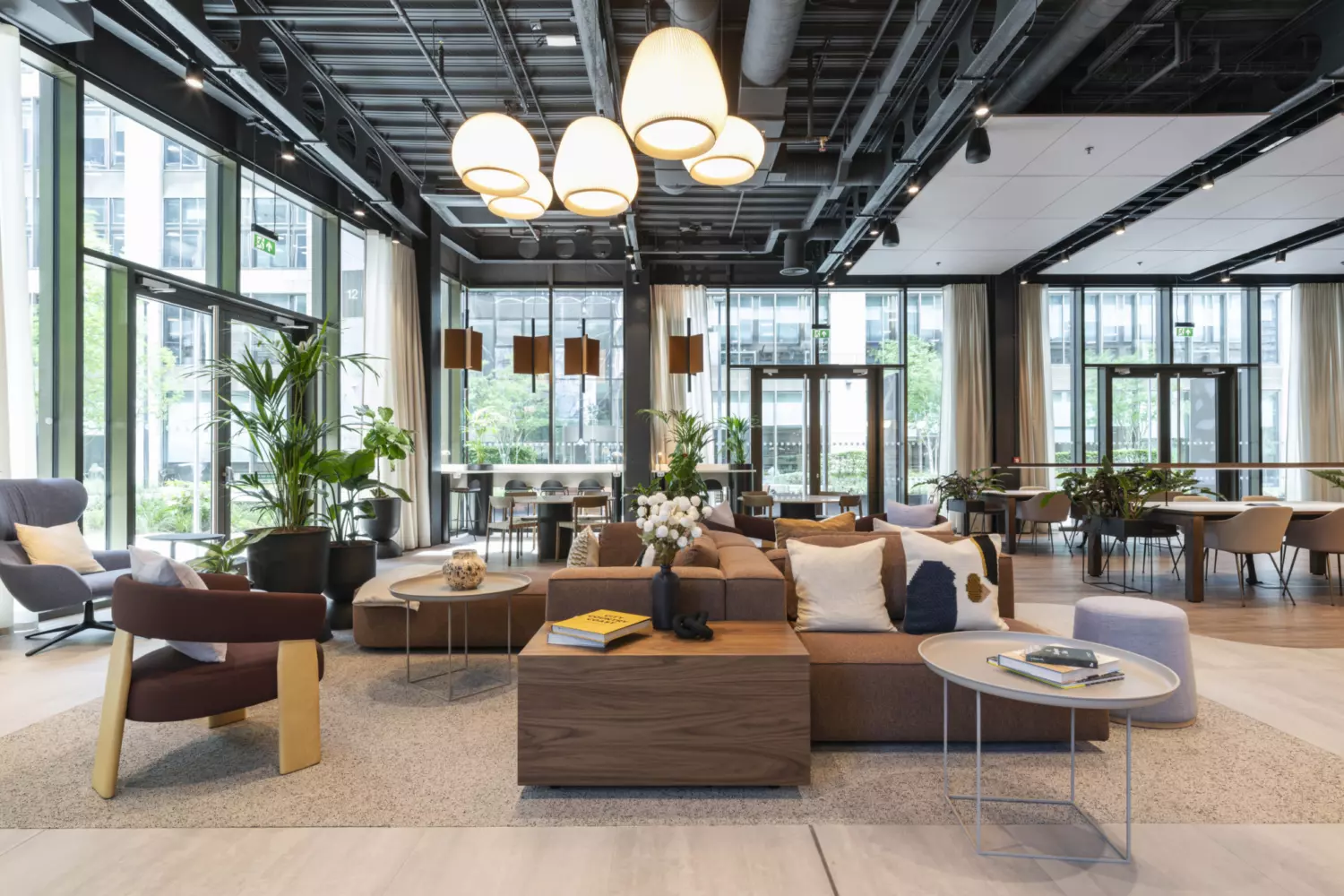 Modern office lounge area featuring a mix of contemporary furniture, including modular sofas, armchairs, and round coffee tables arranged on a textured rug. The space has large floor-to-ceiling windows for natural light, complemented by pendant lighting and exposed ceiling elements for an industrial aesthetic. Indoor plants add greenery, while neutral tones with wood accents create a warm, inviting atmosphere suitable for collaborative work and informal meetings.