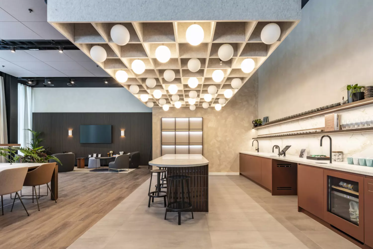 Contemporary office kitchen and breakout area featuring a central island with bar stools, a grid style ceiling with spherical pendant lights and a sleek counter with integrated appliances and shelving. Warm wood tones and neutral finishes create a sophisticated look, while the open plan layout connects to a lounge space with soft seating and a wall mounted screen, ideal for informal meetings and social interaction.