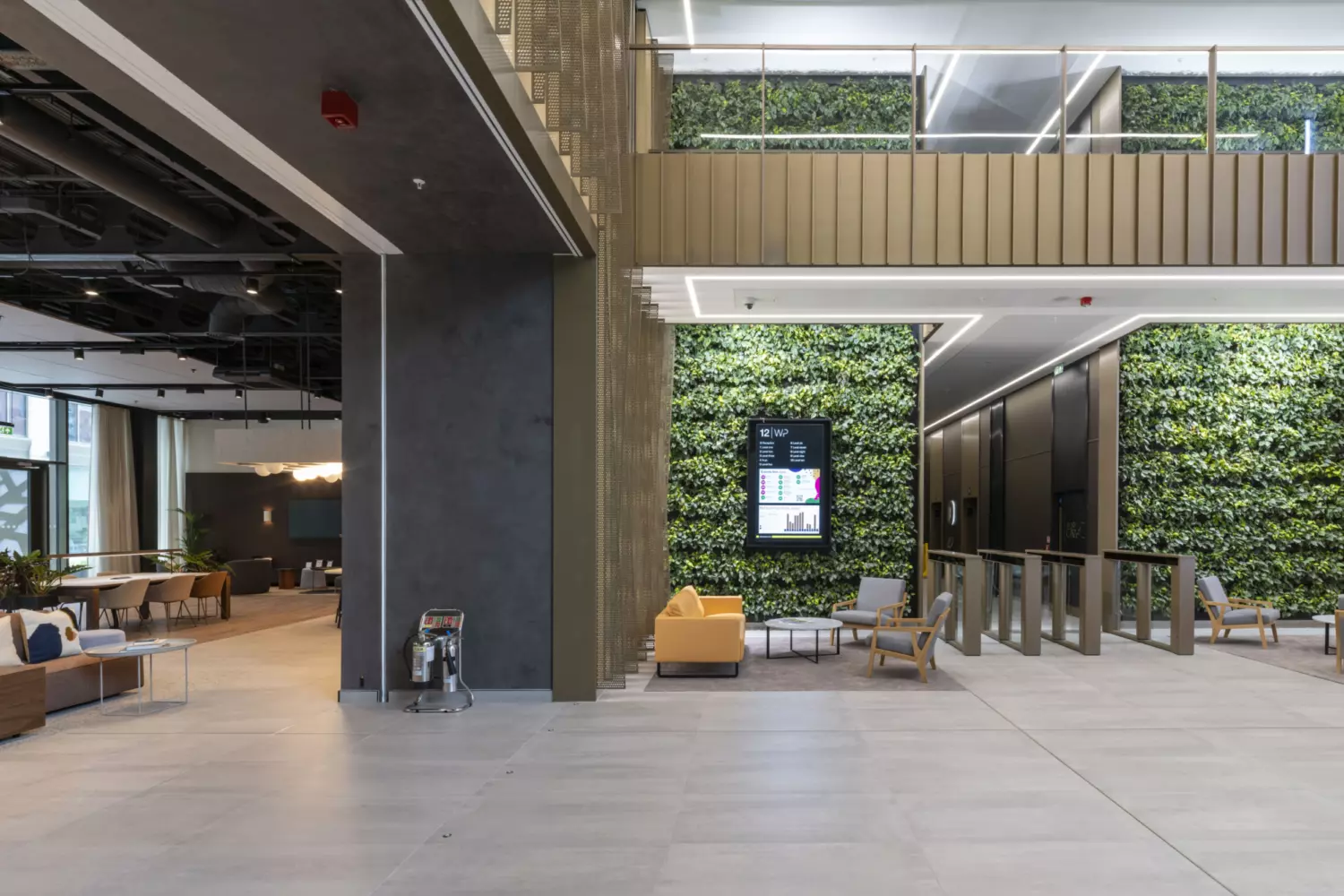 Spacious office reception and lounge area featuring a double height space with living green walls, modern seating and a digital display mounted on a vertical garden backdrop. The design includes sleek turnstiles for secure access, a mezzanine level with metal balustrades and an open plan layout with neutral flooring and exposed ceiling elements. The biophilic design and contemporary finishes create a welcoming and sustainable workplace environment.