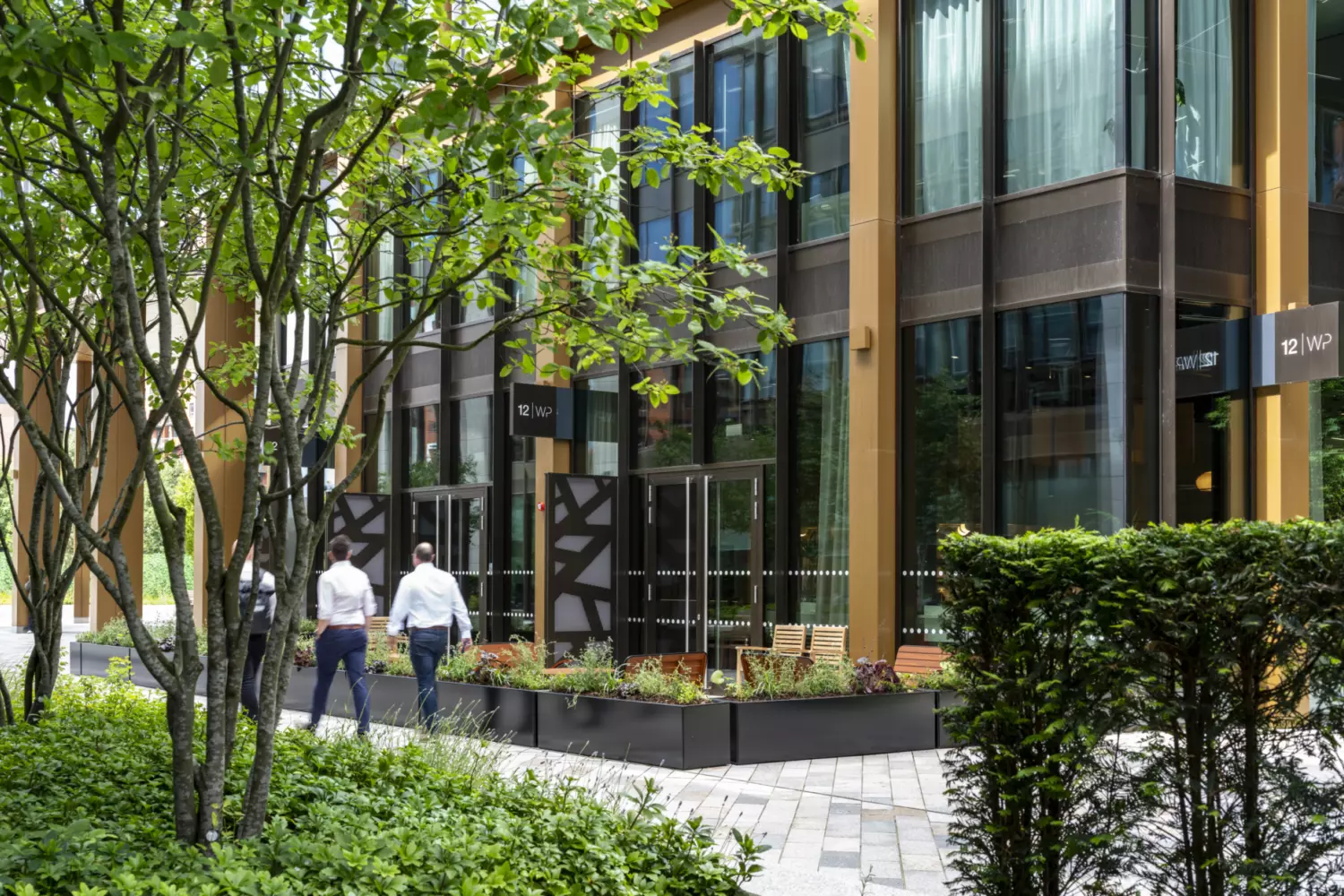 Exterior view of a modern office building with tall glass windows framed by bronze coloured structural elements. The landscaped walkway features lush greenery, trees and raised planters with flowers, creating a welcoming outdoor space. Two people are walking along the paved path, which leads to entrances with contemporary design details, reflecting a high quality office fit out and biophilic design approach.