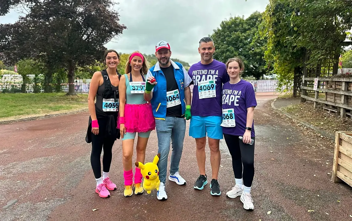 Five participants in colourful fancy dress stand together on a road at an organised running event. One person is dressed as a Pokémon character, wearing a blue vest, red-and-white cap and green gloves, with a Pikachu toy at their feet. Another wears a bright pink tutu and matching accessories, while two participants wear purple charity event T-shirts. All five are wearing race bibs. Trees, fencing and an overcast sky form the background.