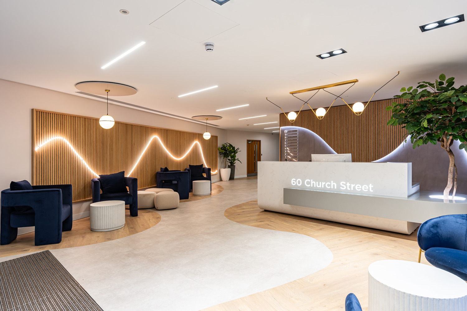 A modern office reception area with a clean, minimalist design. The space features a light wood and white colour scheme with a wave-like LED light pattern on a slatted wooden wall. The reception desk is white with "60 Church Street" lit up on the front. There are navy blue armchairs, round pouffes and small white tables arranged in seating areas. Large potted plants add greenery and contemporary pendant lights hang from the ceiling. The flooring blends light wood and white finishes in a flowing pattern.